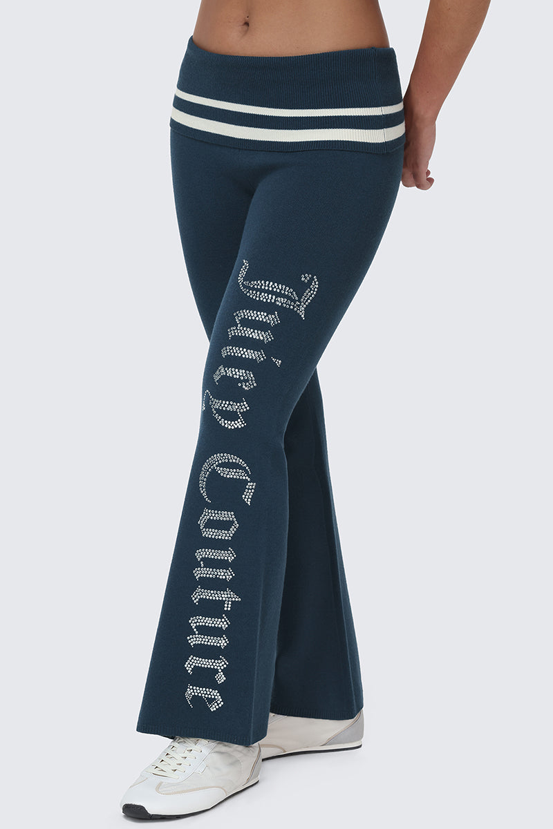 A person is wearing the Big Bling Knit Flare Pants—blue sporty flared pants featuring white stripes at the waistband and "Juicy Couture" in rhinestones down the leg—paired with white sneakers, standing against a plain light background.