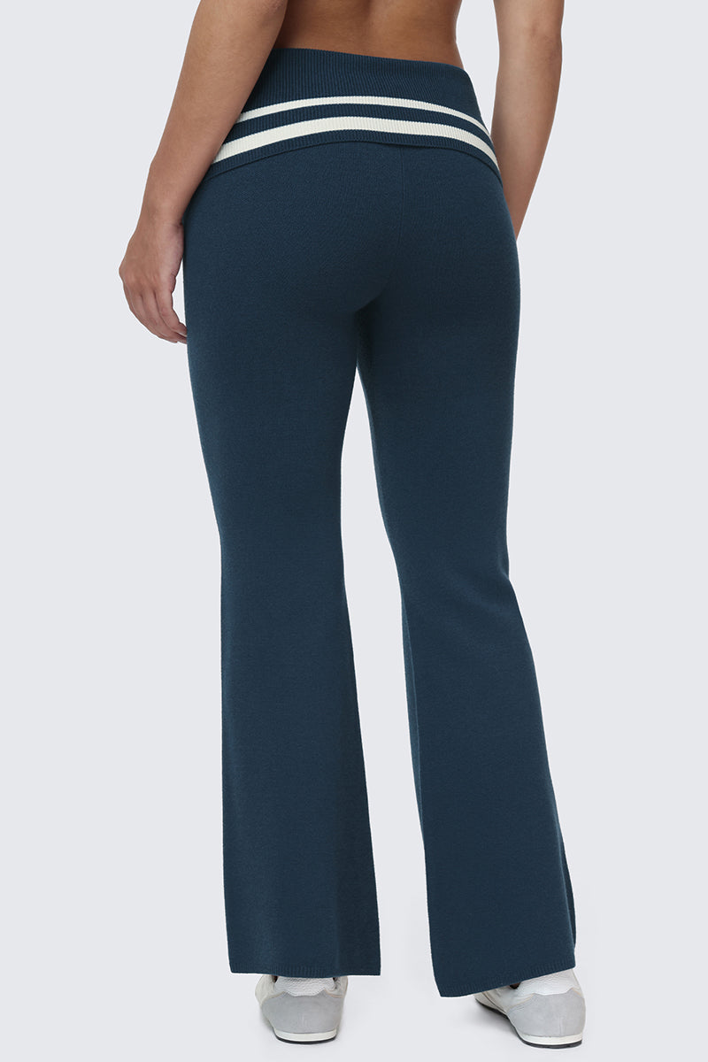 A person wearing the Big Bling Knit Flare Pants—high-waisted, dark blue sporty flare pants featuring two white stripes at the waistband—paired with white sneakers and standing against a light gray background.