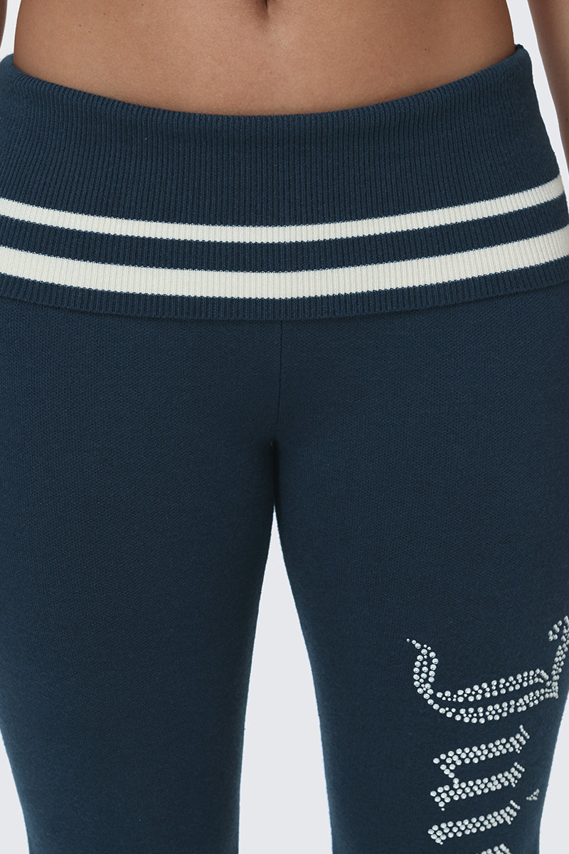 Close-up of a person wearing the Big Bling Knit Flare Pants—high-waisted, dark blue sporty flare pants featuring two white stripes on the waistband and sparkling rhinestone-like embellishments on the left thigh.