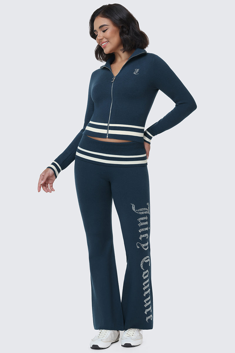 A woman is wearing the Big Bling Knit Flare Pants, a navy blue zip-up tracksuit with white stripes on the cuffs and waist. The sporty flare pants feature "Juicy Couture" written in white script down one leg. She is smiling and looking to the side.