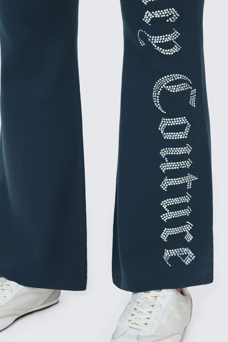 A close-up of a person wearing the Big Bling Knit Flare Pants in dark blue, featuring Juicy Couture in rhinestones on the leg, paired with white sneakers.