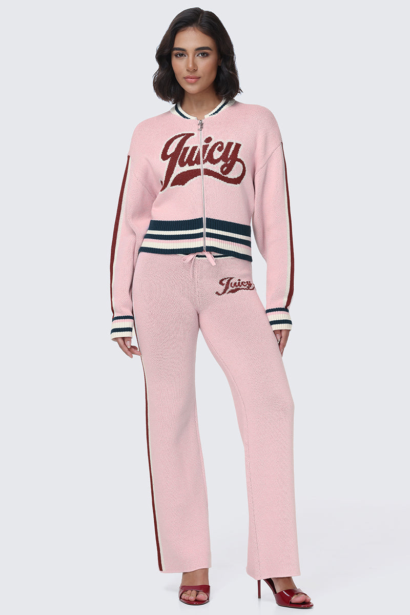 A woman stands against a plain light background, wearing red open-toe heels and a pink knit tracksuit featuring the Juicy script logo on the top and pants. The look is styled with Contrast Knit Wide Leg Pants to complete this sporty essential with an effortlessly chic touch.