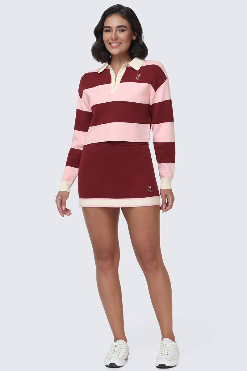 A woman is wearing the Small Bling Knit Cropped Polo—a maroon and pink striped long-sleeve knit polo rugby shirt with a cropped silhouette—paired with a maroon mini skirt and white sneakers, standing and smiling against a plain light background.