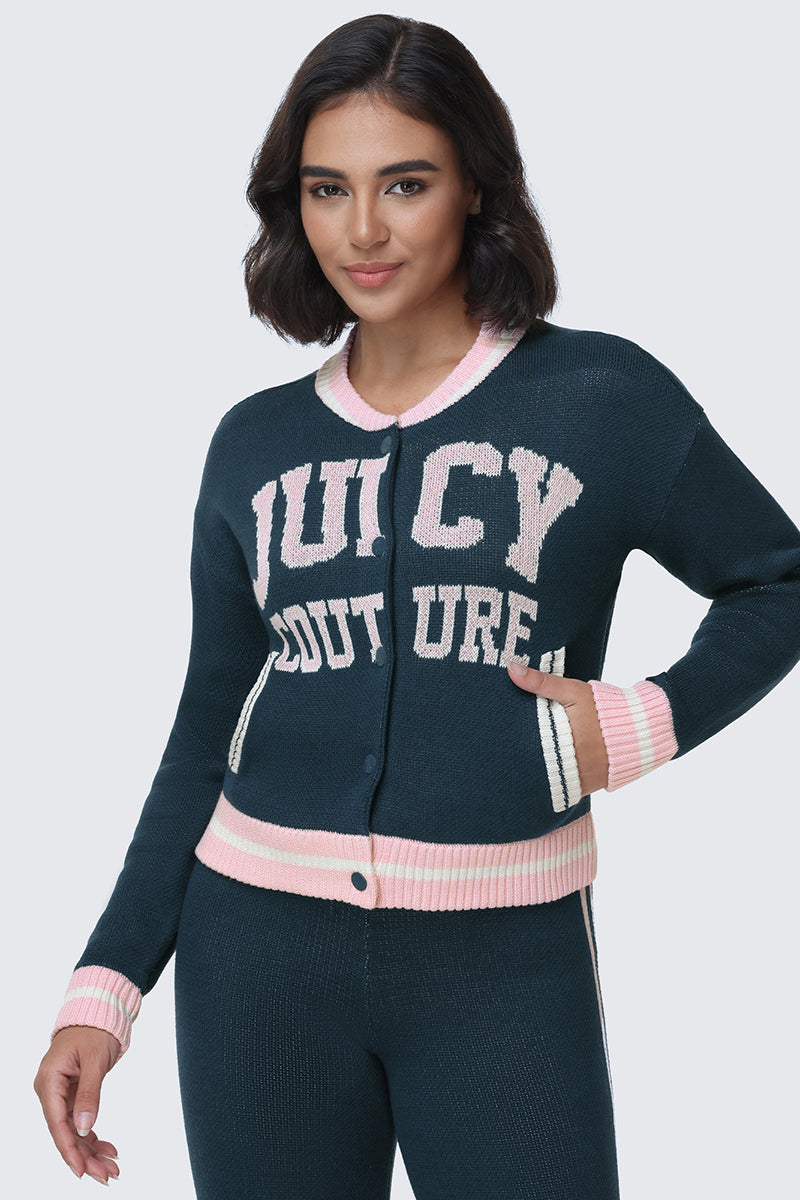A woman wearing the Collegiate Knit Bomber Jacket by Juicy Couture in navy blue with pink and white trim, along with matching pants, poses with one hand on her hip and a subtle smile. The knit bomber features bold “JUICY COUTURE” lettering across the front.