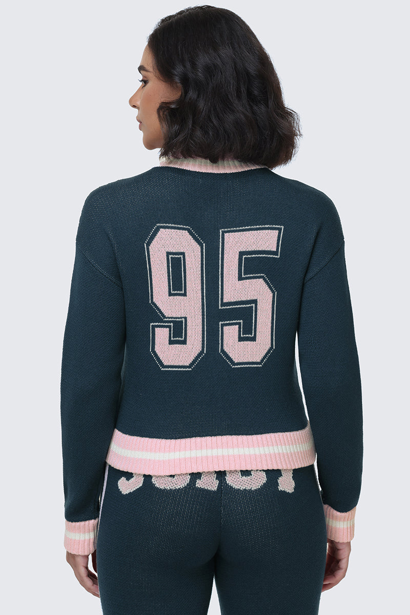 A woman with shoulder-length dark hair is shown from the back, wearing a dark Collegiate Knit Bomber Jacket by Juicy Couture featuring a pink "95" on the back, paired with matching pants with pink trim and text.