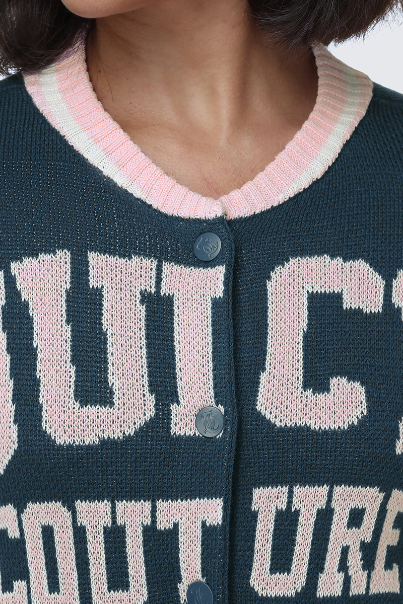 A person is wearing the Collegiate Knit Bomber Jacket in dark green by Juicy Couture, which features large pink and white letters, a pink and white ribbed collar, and dark buttons. Only the lower face, neck, and upper torso are visible.