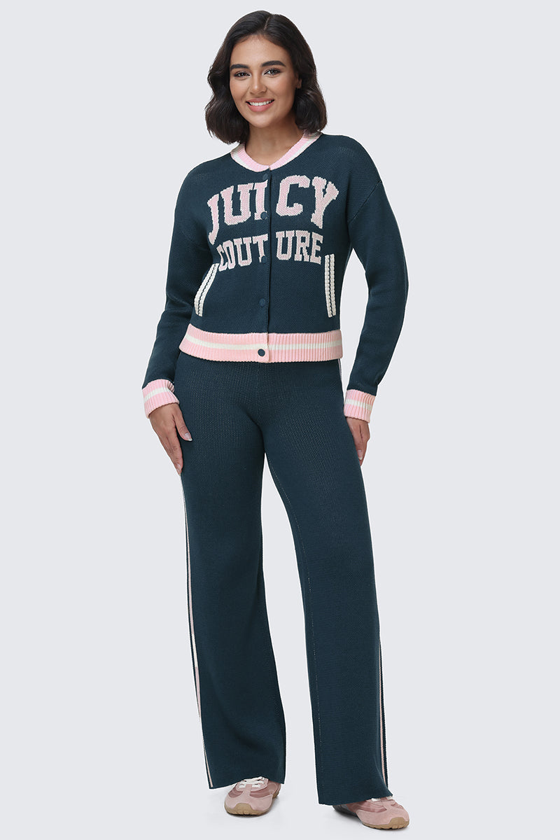 A woman stands and smiles, wearing a dark blue Juicy Couture Collegiate Knit Bomber Jacket with matching pants featuring pink and white accents. The varsity jacket displays JUICY COUTURE across the chest, and she completes the look with pink shoes.
