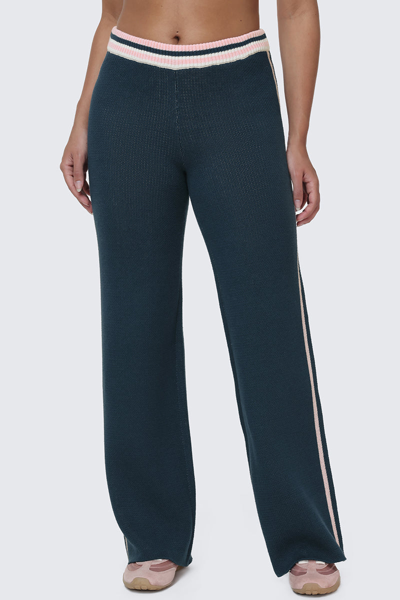 Wearing the Collegiate Knit Wide Leg Pants—high-waisted, dark teal knit with pink and white striped accents on the waistband and sides, plus subtle logo detailing for a varsity-inspired touch—paired with light pink sneakers.