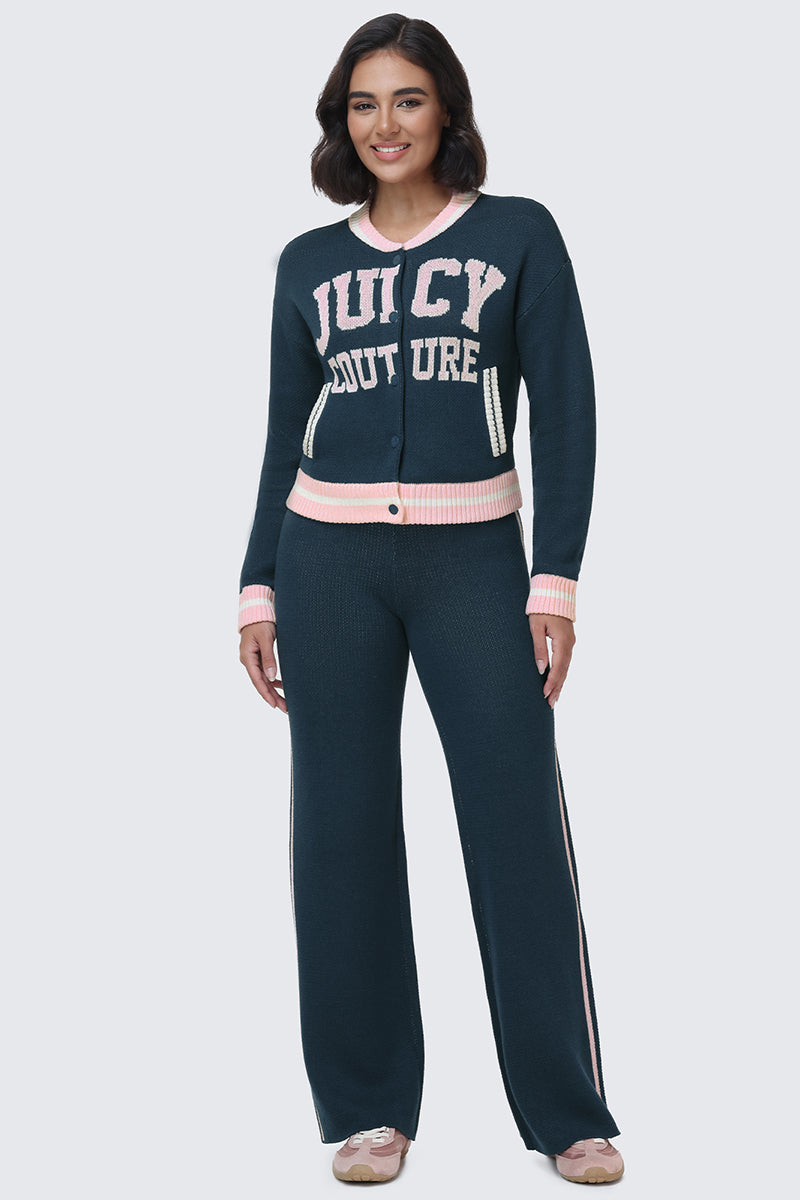 A woman stands smiling, wearing a matching dark blue knit set. Her outfit features the Collegiate Knit Wide Leg Pants, which have bold pink stripes, paired with a varsity-style sweater displaying large pink and white JUICY COUTURE logo detailing along with pink trim on the collar and cuffs. She completes the look with beige shoes.