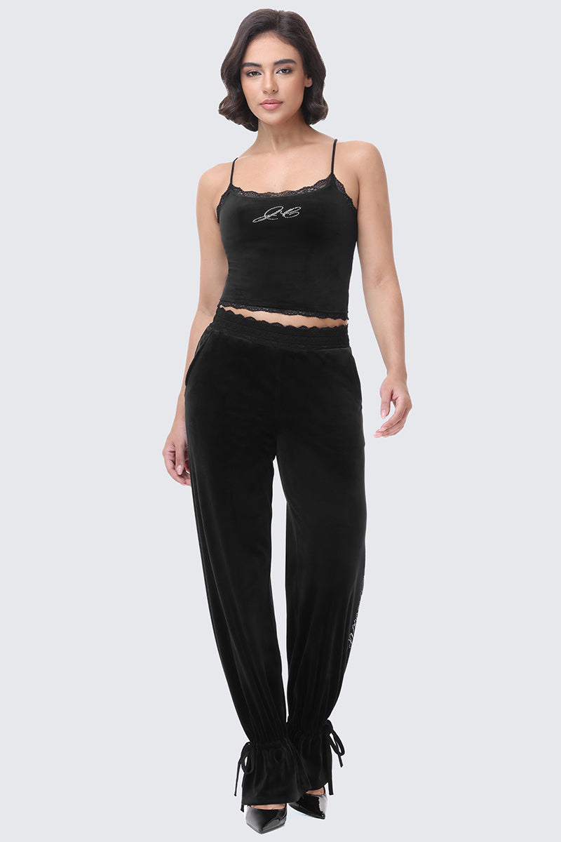 A woman wears a black Small Bling Cropped Cami with lace trim and rhinestone lettering, paired with matching high-waisted lounge pants featuring a lace waistband and drawstring cuffs.