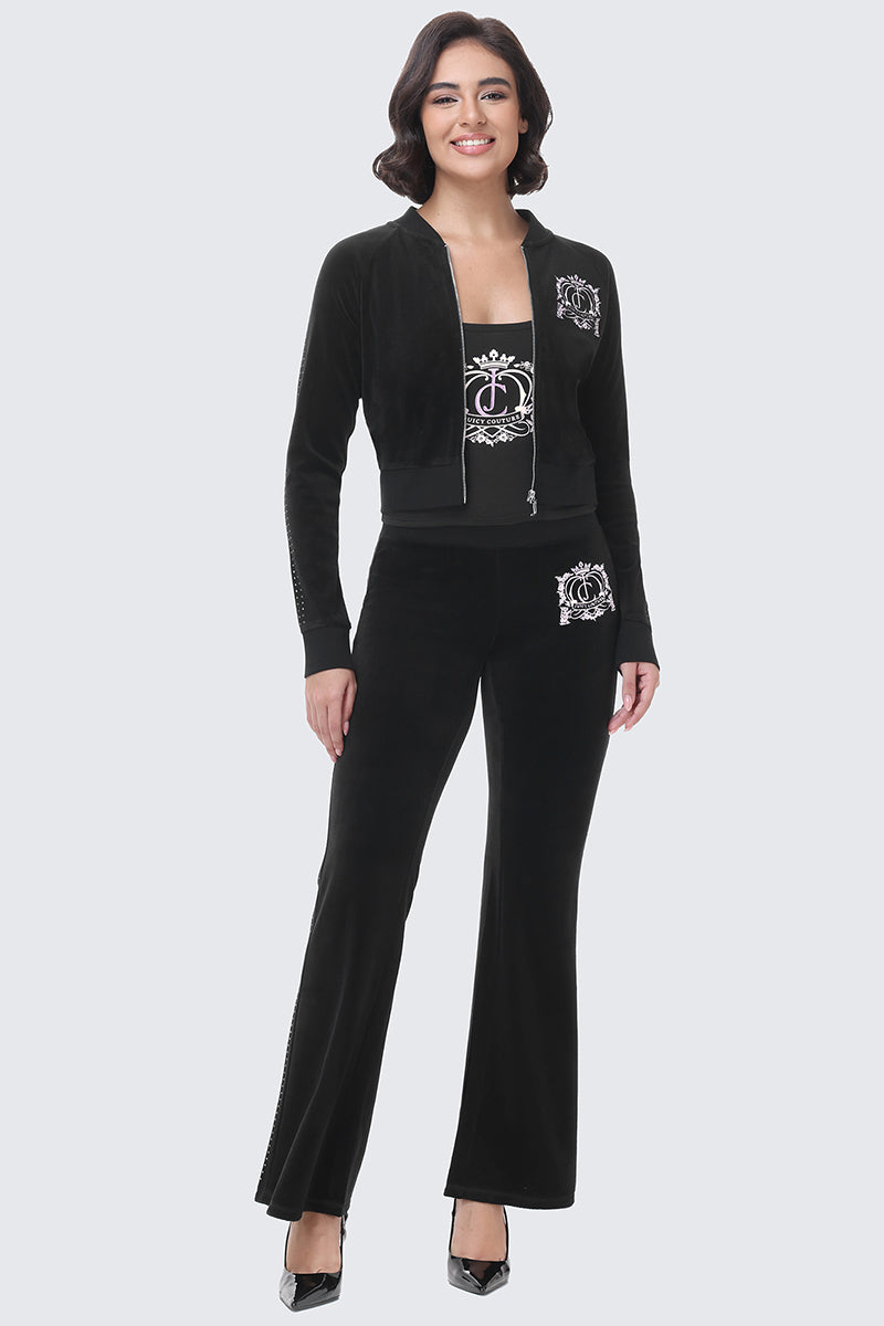 A smiling model wears a black cotton velour Scottie Crown Cotton Velour Jacket and matching flared pants, creating a sporty silhouette with a decorative white crest graphic on the chest and hip.