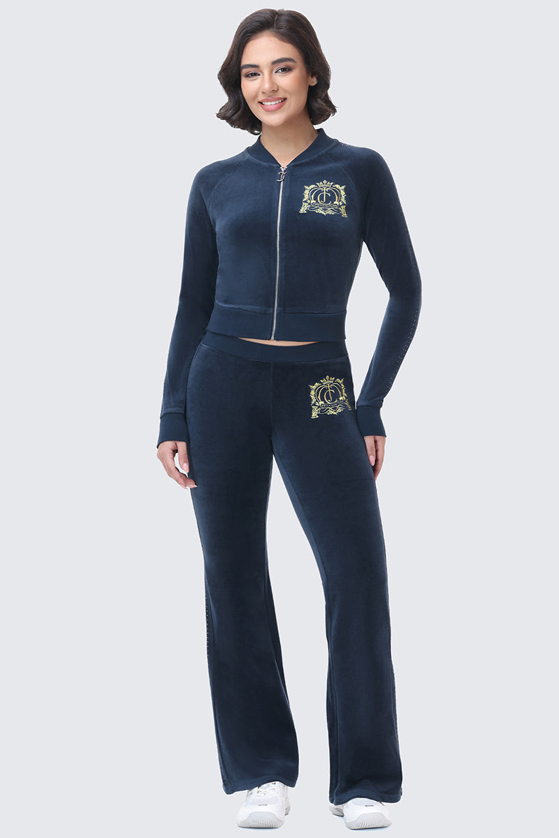 A woman wears a Scottie Crown Cotton Velour Jacket and matching flared pants, both featuring an embroidered gold crest logo on the chest and hip.
