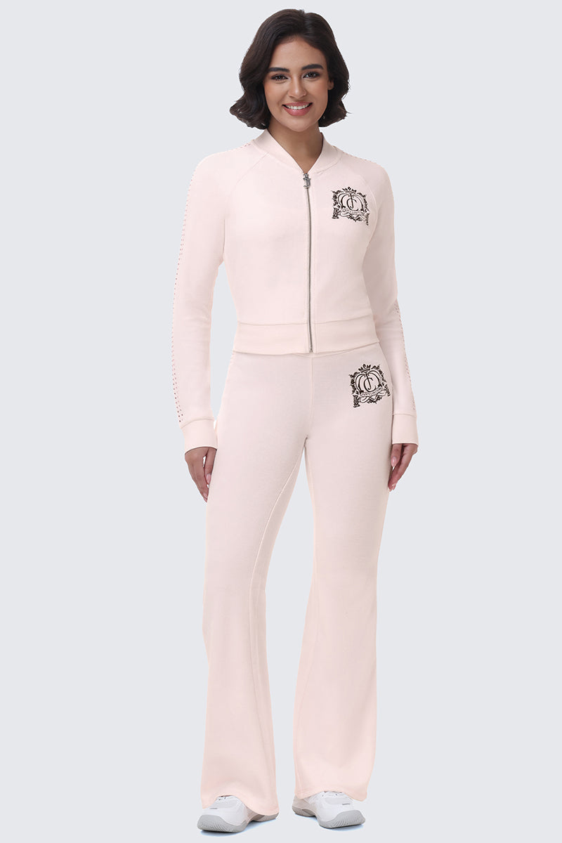 A smiling woman wears a pale pink Scottie Crown Cotton Velour Jacket and matching flared pants, both featuring a crest logo on the left side.
