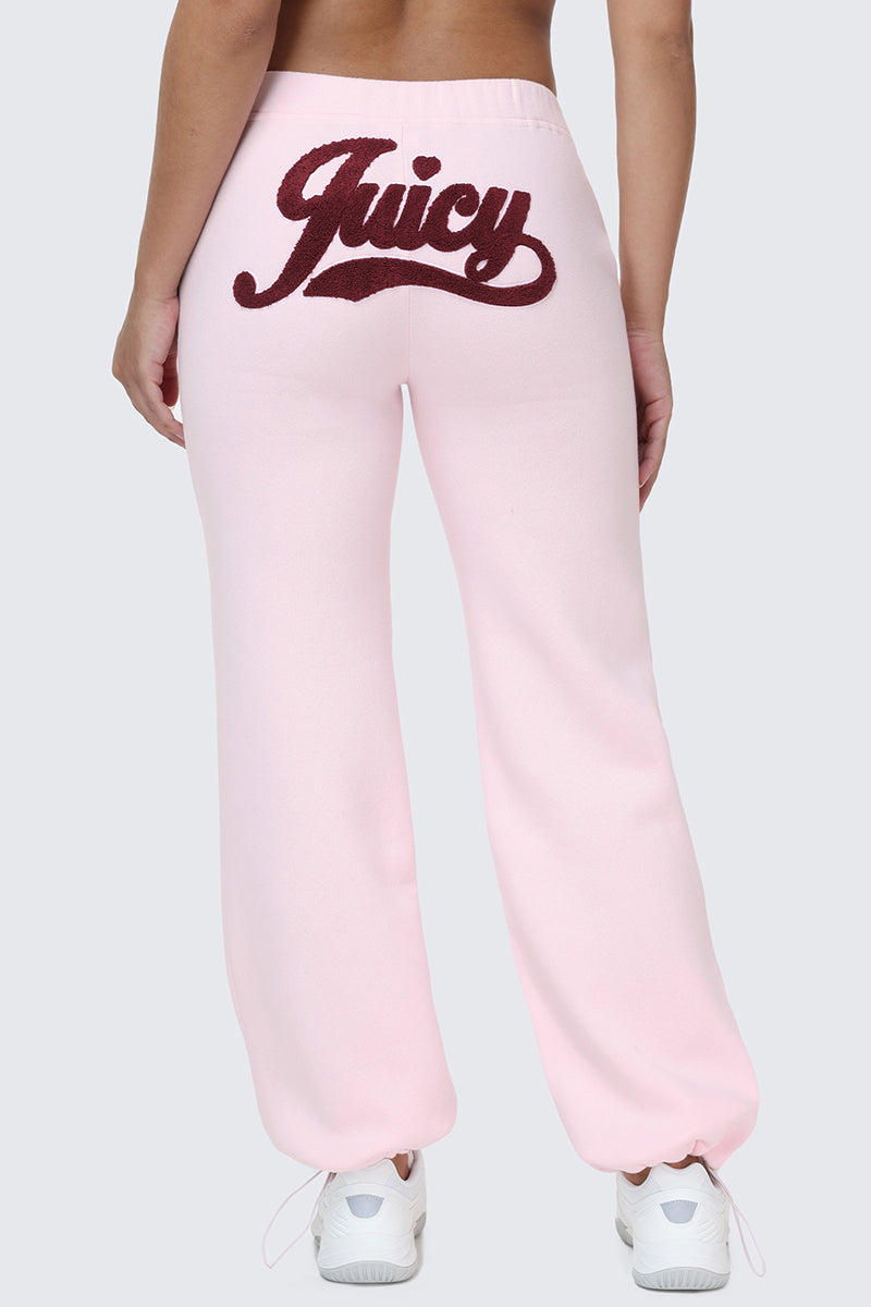 A person wearing the Embroidered Knit Bubble Leg Pants in light pink, featuring the word "Juicy" in large, dark red script across the back, paired with white sneakers and shown from the waist down, viewed from behind.