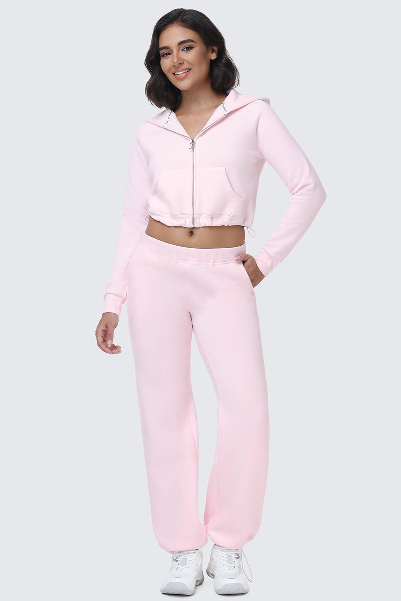 A woman stands and smiles with one hand in her pocket against a plain light gray background, wearing a light pink Juicy zip-up hoodie and matching Embroidered Knit Bubble Leg Pants.