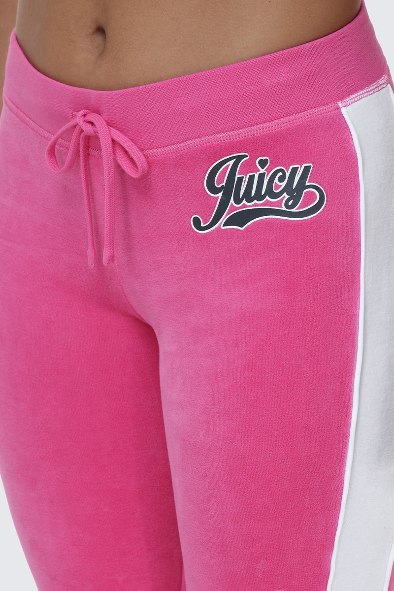 A close-up of a person wearing Cotton Velour Track Pants in bright pink, featuring a white side stripe and bold black "Juicy" script on the left hip, finished with stylish flap back pockets that create an effortlessly off-duty look.
