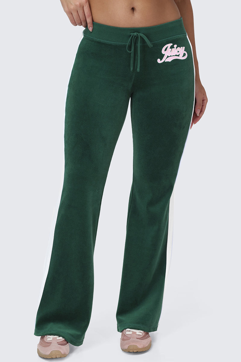 Wearing the Cotton Velour Track Pants, which feature a green velour fabric, white side stripes, a drawstring waist, flap back pockets, and the word "Juicy" in white script on the left hip, this person pairs them with light pink sneakers.