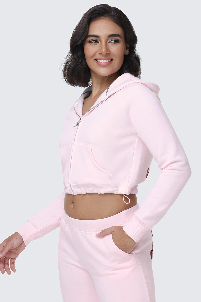 A woman wearing the Embroidered Knit Cropped Hoodie in light pink with matching sweatpants smiles while looking to the side, one hand in her pocket.