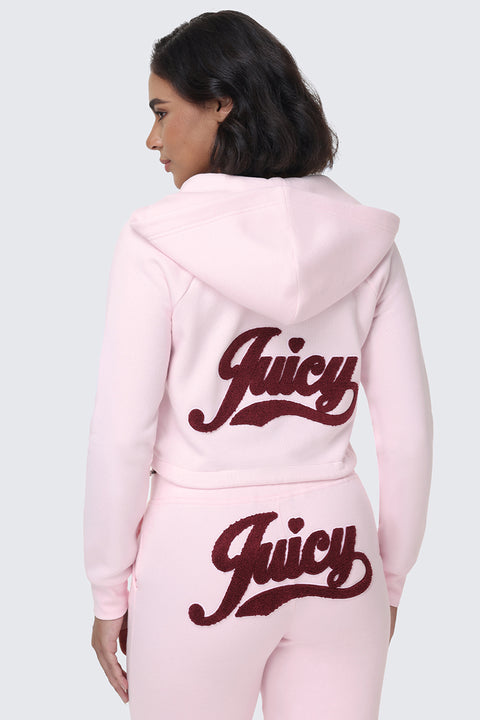 A woman stands against a plain gray background wearing the Embroidered Knit Cropped Hoodie in light pink, paired with matching sweatpants. Both pieces feature the word "Juicy" in large maroon letters on the back and bottom, while the embroidered logo adds a stylish touch to this coordinated look.