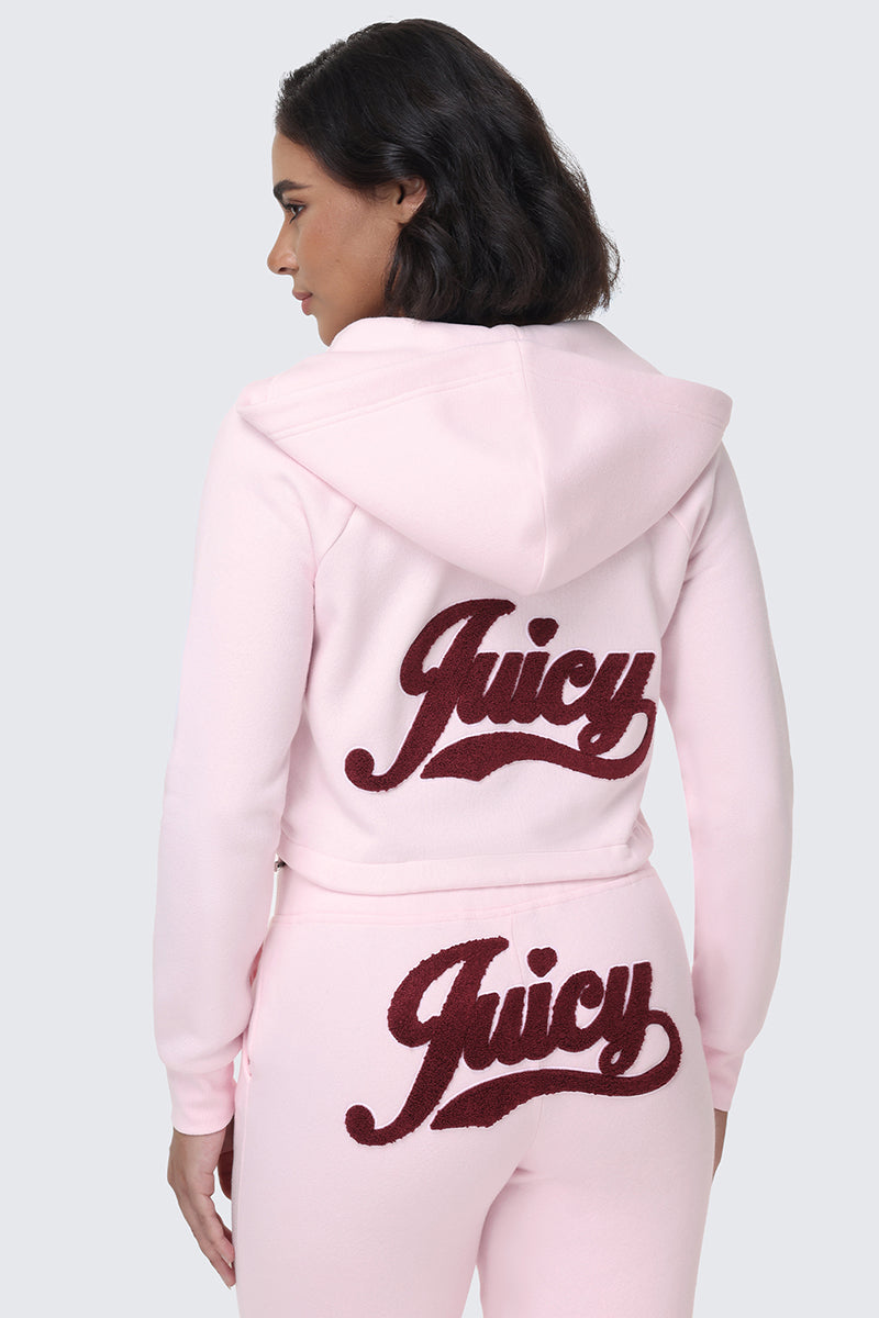 A woman stands against a plain gray background wearing the Embroidered Knit Cropped Hoodie in light pink, paired with matching sweatpants. Both pieces feature the word "Juicy" in large maroon letters on the back and bottom, while the embroidered logo adds a stylish touch to this coordinated look.