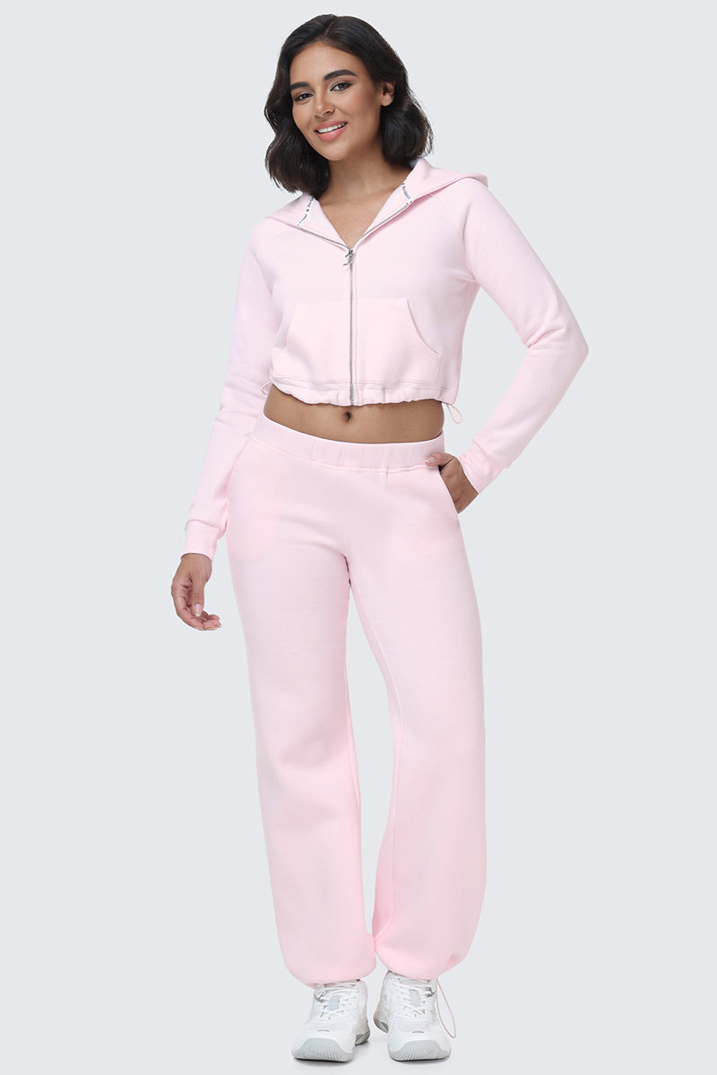 A woman in an Embroidered Knit Cropped Hoodie, light pink in color with a zip front and embroidered logo, pairs it with matching Juicy Couture sweatpants and white sneakers. She stands against a plain light gray background, smiling with one hand on her hip.