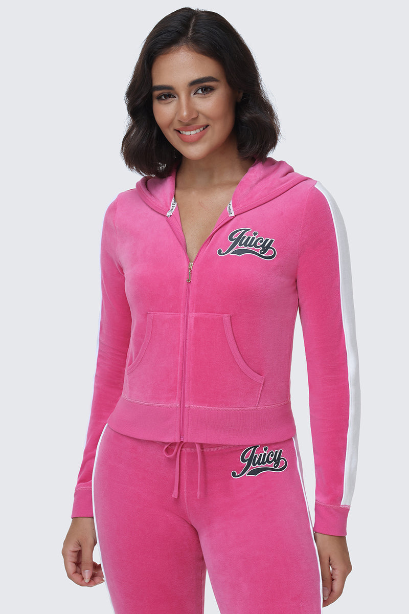 A person stands smiling against a plain light background, dressed in the Cotton Velour Hoodie—a pink zip-up tracksuit featuring sporty white sleeve stripes and the signature "Juicy" script logo on the chest and hip.