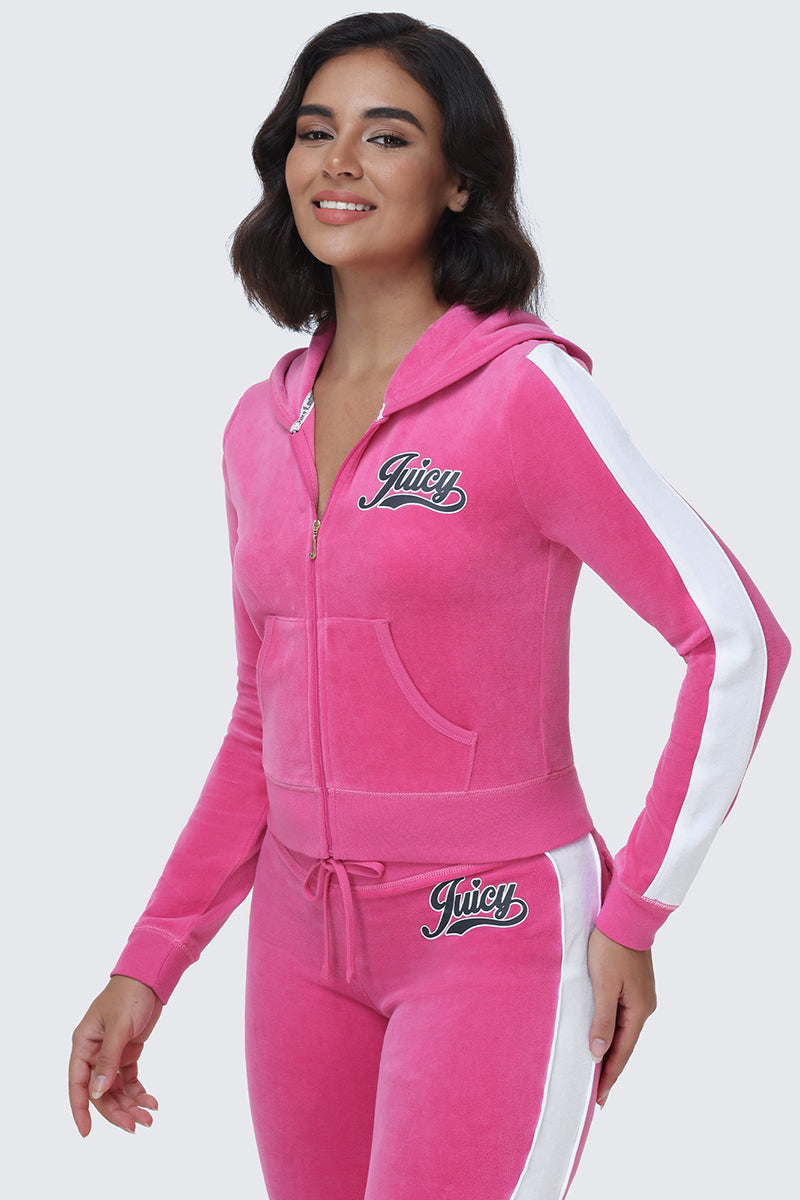 A woman with dark hair smiles as she stands against a light gray background, wearing the Cotton Velour Hoodie in pink. The hoodie features white stripes along the sleeves and legs, and showcases Juicy’s signature script logo on the chest and hip.