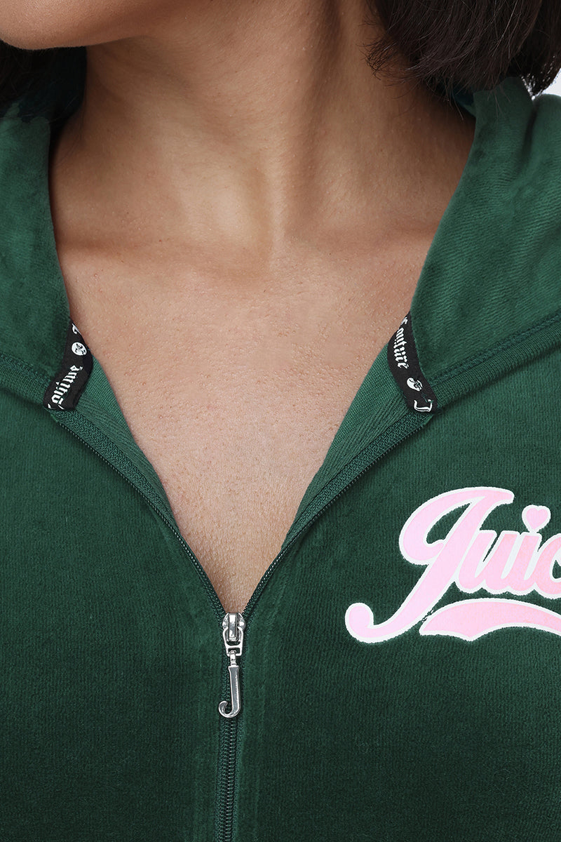 A close-up shows a person wearing the Cotton Velour Hoodie in green, featuring the signature “Juicy” script logo in pink on the left side. With its front zipper partially open to reveal a hint of chest and neck, this sporty hoodie offers a relaxed yet stylish vibe.