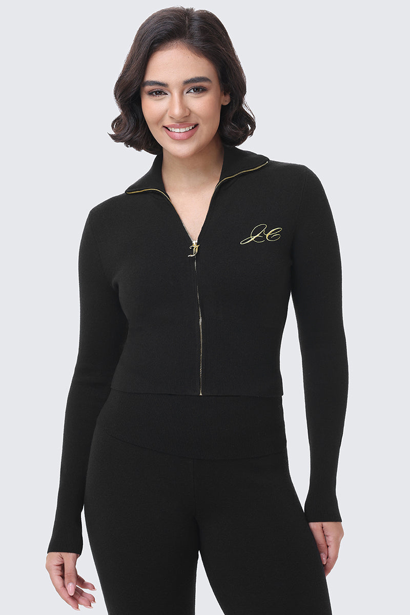 A woman wears a black, long-sleeved, cropped Embroidered Knit Cropped Jacket featuring a gold zipper and a gold embroidered Juicy Couture script logo on the chest.