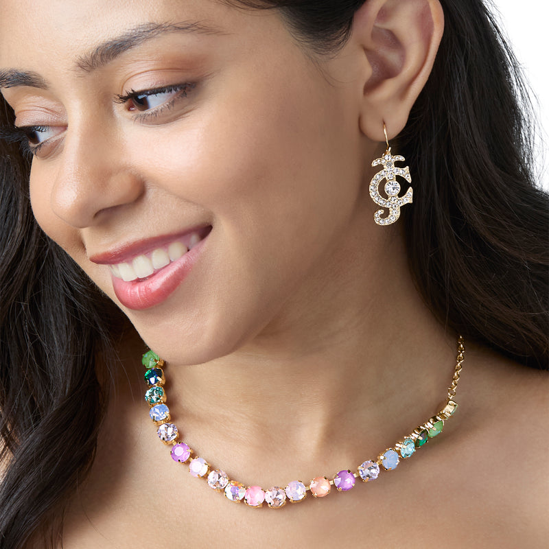 A woman smiles while wearing a gold chain necklace with colorful gemstones and the Rhinestone Logo Drop Earrings from Juicy Couture, featuring interlocking G-shaped logos encrusted with rhinestones for a bold statement jewelry look.