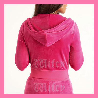 A person shown from the back wearing a bright pink velour hooded sweatshirt and matching bottoms, with the name 'Wifey' in a gothic font across the back and waistband. The photo is framed by a pink border on a white background.