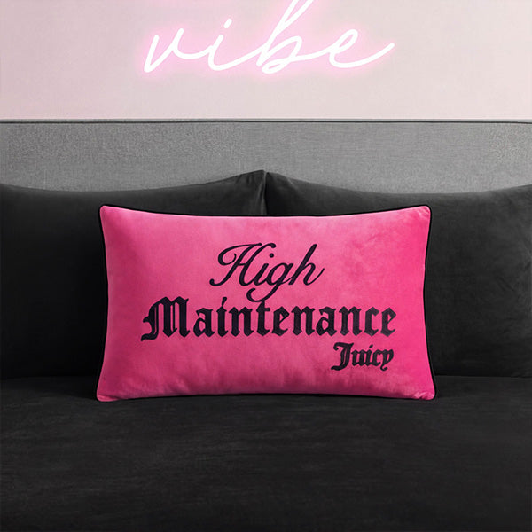 A bright pink rectangular throw pillow with black gothic-style text reading 'High Maintenance Juicy' resting on a dark gray couch, with a pink neon-style 'vibe' sign above.