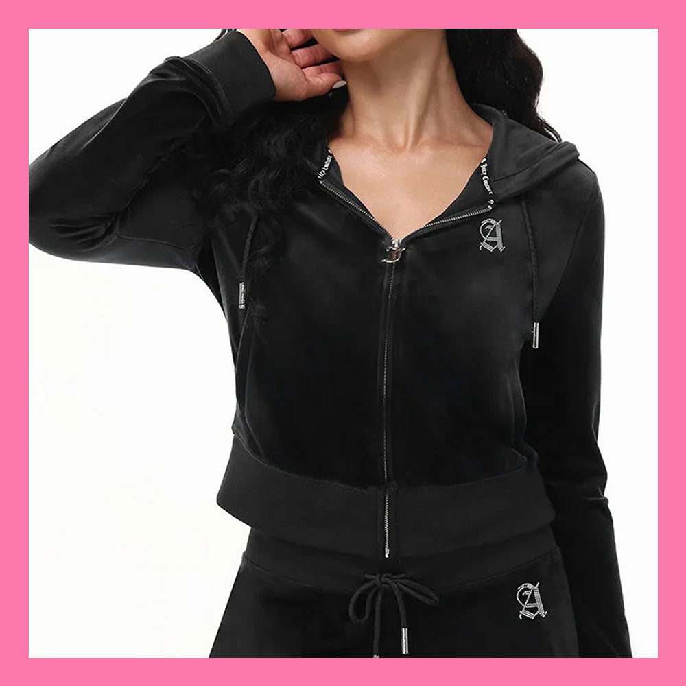 A woman wearing a black zip-up hooded sweatshirt and matching black drawstring bottoms, shown from the neck down against a white background with a pink border. The outfit has a small white embroidered letter 'A' logo on the chest and waist.