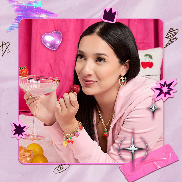 Collage-style image of woman in pink top wearing gold earrings, a necklace, and bracelet with cherry charms, holding a cocktail glass. Y2K and scribble-style stickers and graphics surround the image.