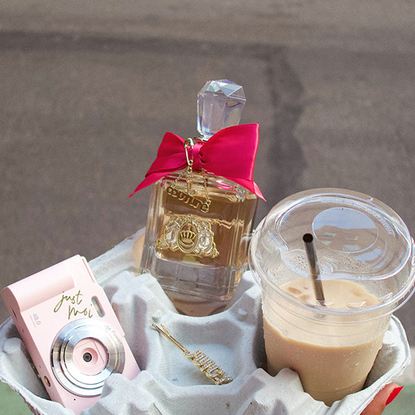 A styled product shot featuring a clear perfume bottle with a bright pink bow and a gold 'COUTURE' emblem, alongside a pink 'Just Moi!' camera, a gold bobby pin, and a plastic cup of iced coffee in a white paper tray.
