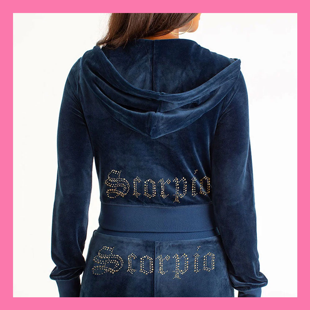 A person shown from the back wearing a navy blue velvet hooded sweatshirt and matching bottoms, both featuring 'Scorpio' spelled out in rhinestones across the back. The image is framed with a pink border.