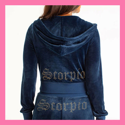 A person shown from the back wearing a navy blue velvet hooded sweatshirt and matching bottoms, both featuring 'Scorpio' spelled out in rhinestones across the back. The image is framed with a pink border.