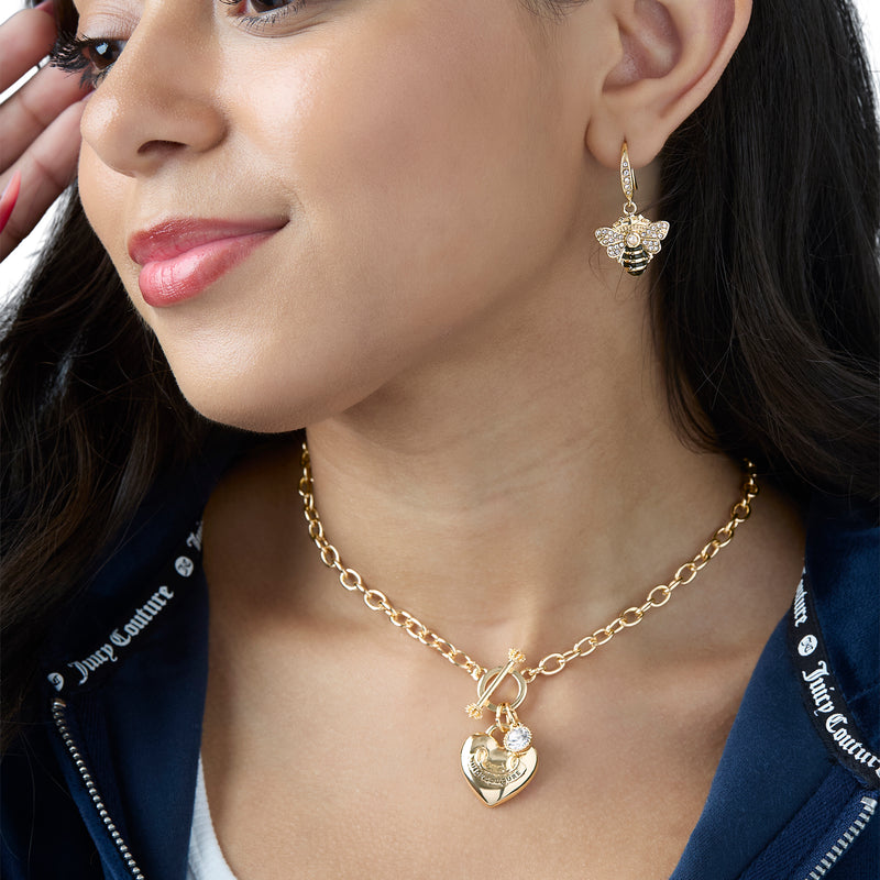 A woman wearing a gold heart pendant necklace and statement queen bee jewelry, featuring Rhinestone and Black Enamel Bee Earrings. She has long dark hair, red lipstick, and is dressed in a navy blue Juicy Couture outfit.