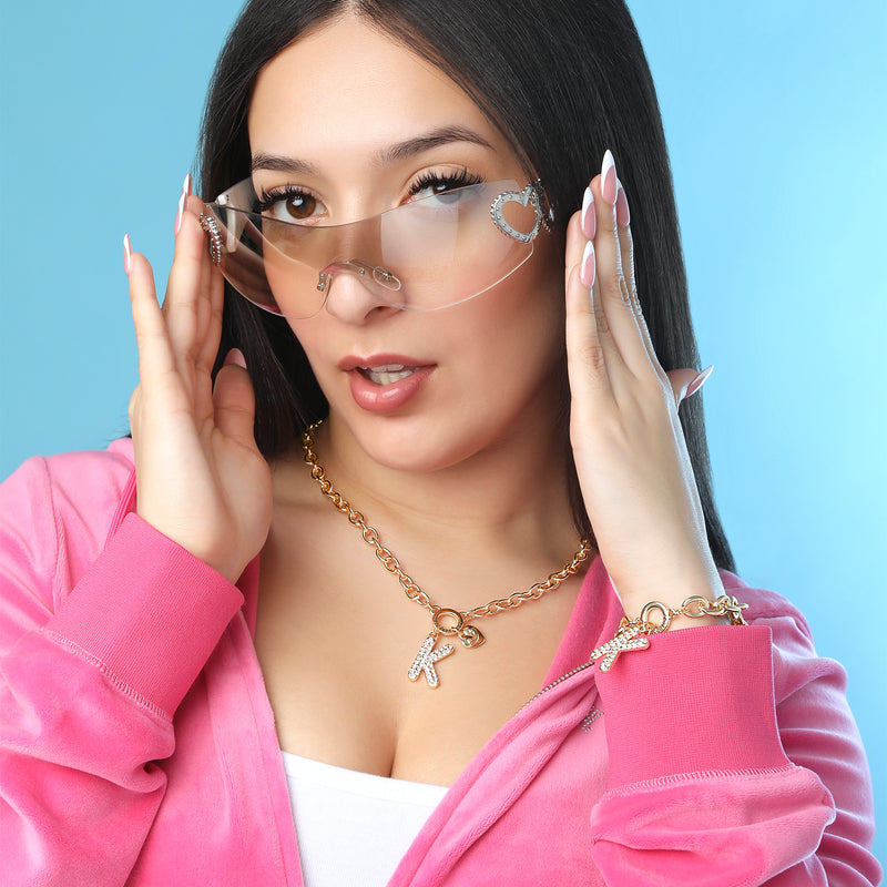 A woman with long dark hair wears a pink velour zip-up hoodie, heart-accented rimless sunglasses, and Y2K-inspired accessories including the Bubble Initial Necklace, Letter K. She poses with her hands near her face against a vibrant blue background.