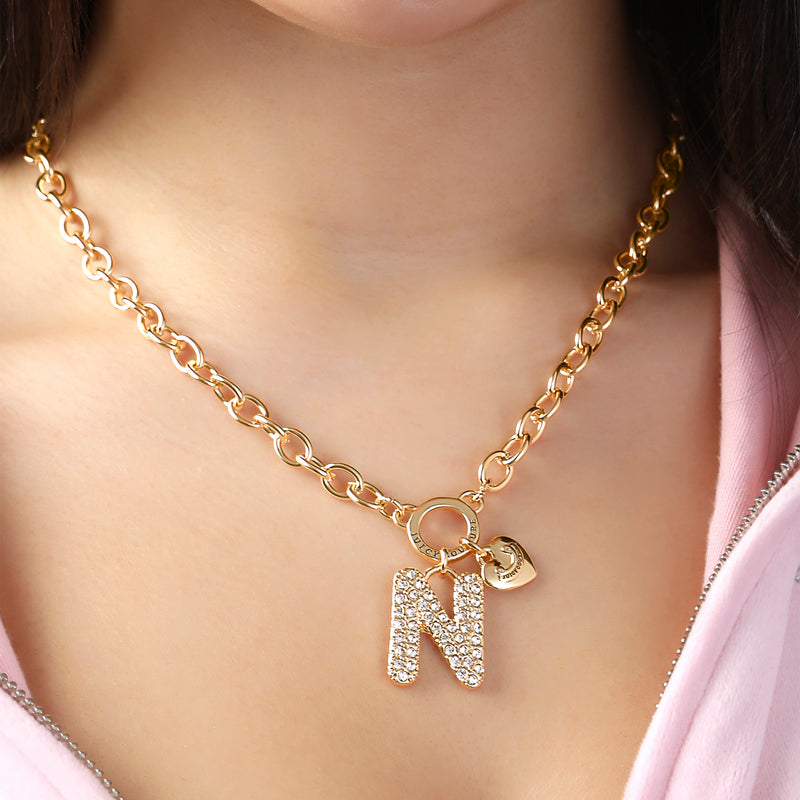 A person wearing the Juicy Couture Bubble Initial Necklace, Letter N—a gold necklace featuring a large, rhinestone-encrusted N initial charm and a small heart charm with the Juicy Couture logo—paired with a light pink hooded garment for a playful Y2K-inspired look.