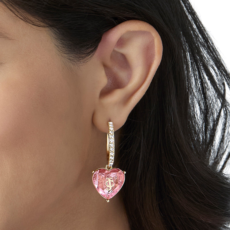 A close-up of a woman’s ear wearing the Heart Charm Hoop Earrings: statement gold hoops adorned with small clear stones and featuring a pink, heart-shaped gemstone pendant—perfect as a heart jewelry gift.