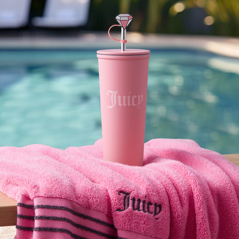 A Couture Hydration Stainless Steel Tumbler Cup from the 6-Piece Set, featuring a pink design with "Juicy" and a diamond straw topper, sits on a matching Juicy towel by the pool with lounge chairs and greenery in the background.