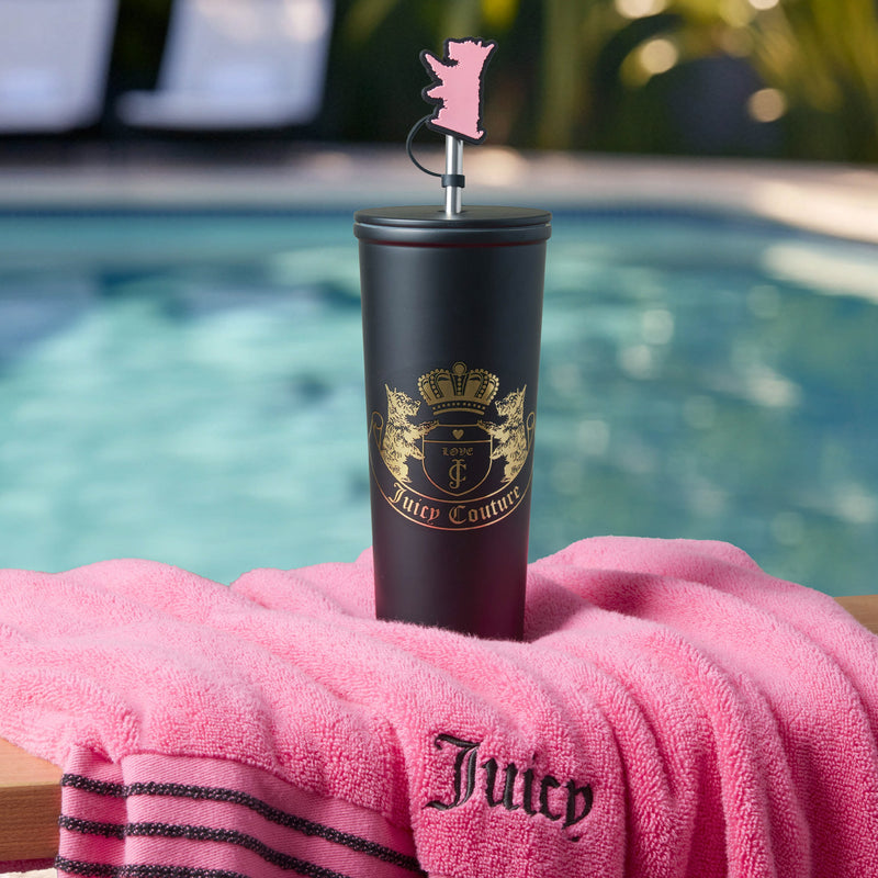 A Couture Hydration Stainless Steel Tumbler Cup from the 6-Piece Set, featuring a gold logo and pink bear topper, rests on a pink Juicy towel by the pool with palm trees and sparkling water in the background.