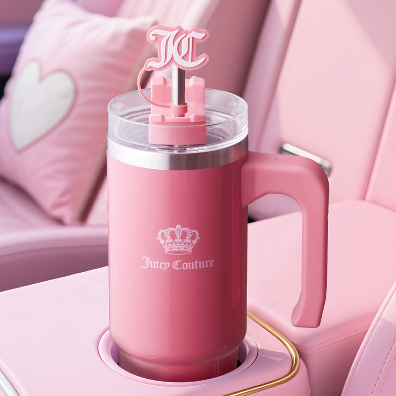 A pink Jetsetter Hydration Stainless Steel Tumbler with handle, clear lid, “JC” logo straw, and neoprene crossbody pouch sits in a pink car’s cup holder with a heart pillow in the background.