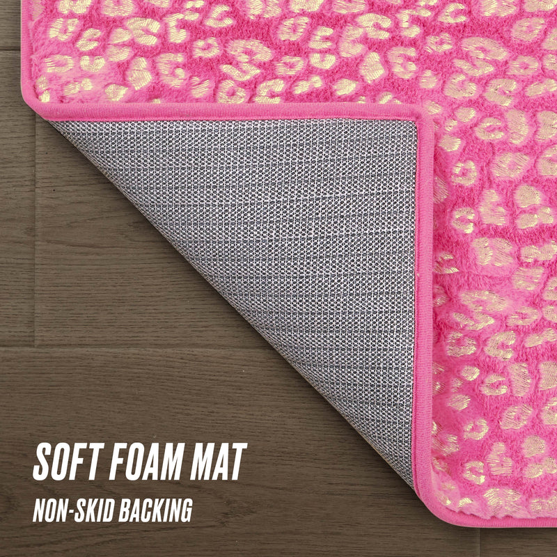 The Metallic Leopard Plush Rug, 2' x 6', features a pink and yellow leopard print with a gray non-skid backing. Shown partially flipped on a wooden floor, the soft foam mat highlights its comfort and secure grip. Text: SOFT FOAM MAT and NON-SKID BACKING.