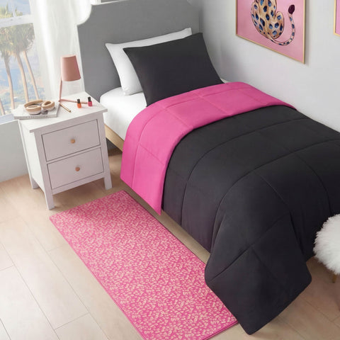 A neatly made twin bed with a black and bright pink comforter, black pillow, and white sheets. A Metallic Leopard Plush Rug, 2' x 6', sits beside the bed near a white nightstand with cosmetics, lamp, and framed leopard artwork.