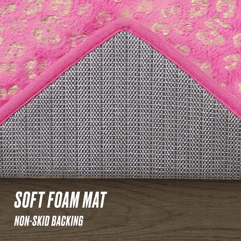 A Metallic Leopard Plush Rug, 3' Heart, with a pink leopard-print soft foam surface is shown on a wood floor. One corner is flipped to show its textured non-skid backing. Text on the image: SOFT FOAM MAT and NON-SKID BACKING.