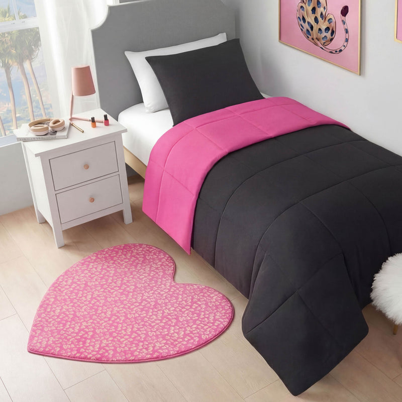 A neatly made bed with a dark gray comforter and bright pink accent sits next to a white nightstand topped with makeup. A Metallic Leopard Plush Rug, 3' Heart, decorates the wooden floor, while a leopard art print hangs on the wall.