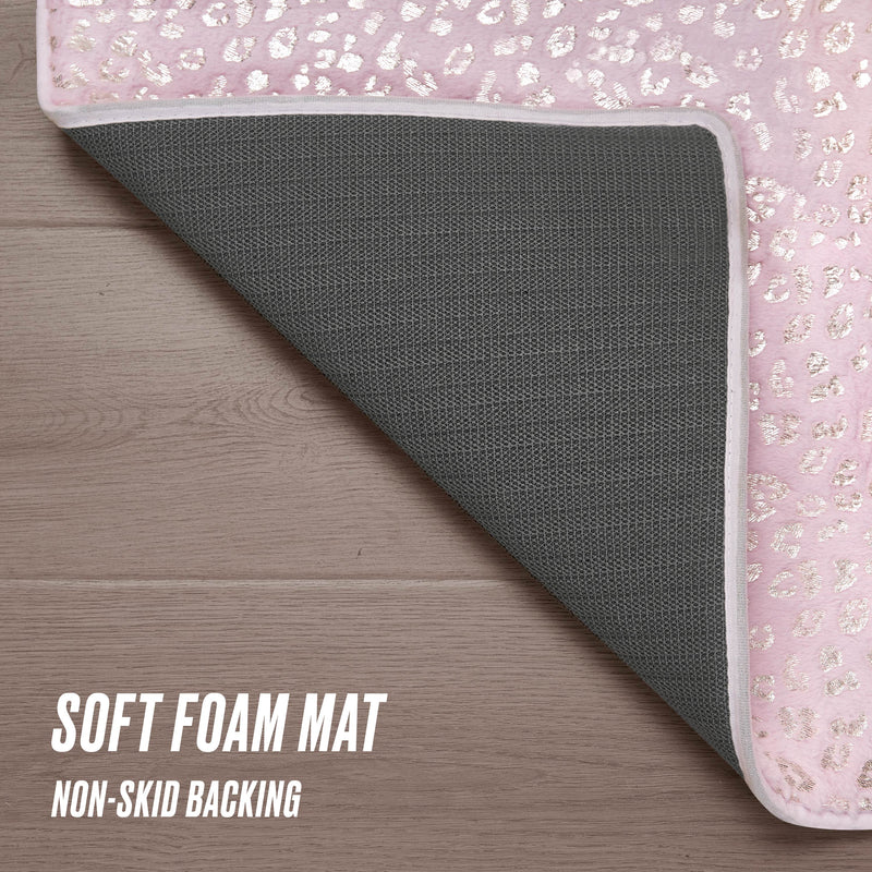 A close-up of the Metallic Leopard Plush Rug, 2' x 6', in pink and white, partially flipped to reveal its gray non-skid backing on a wooden floor. Text: “SOFT FOAM MAT NON-SKID BACKING.”.