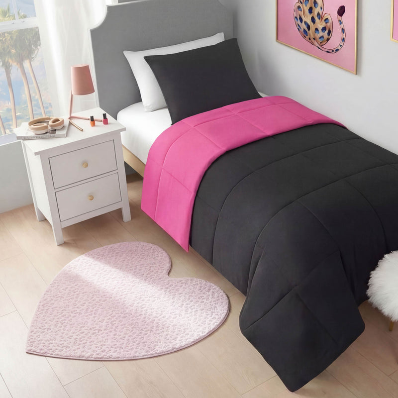 A neatly made bed with a black comforter and bright pink strip sits beside a white nightstand with a lamp, books, and makeup. A Metallic Leopard Plush Rug, 3' Heart, in pink decorates the light wooden floor.